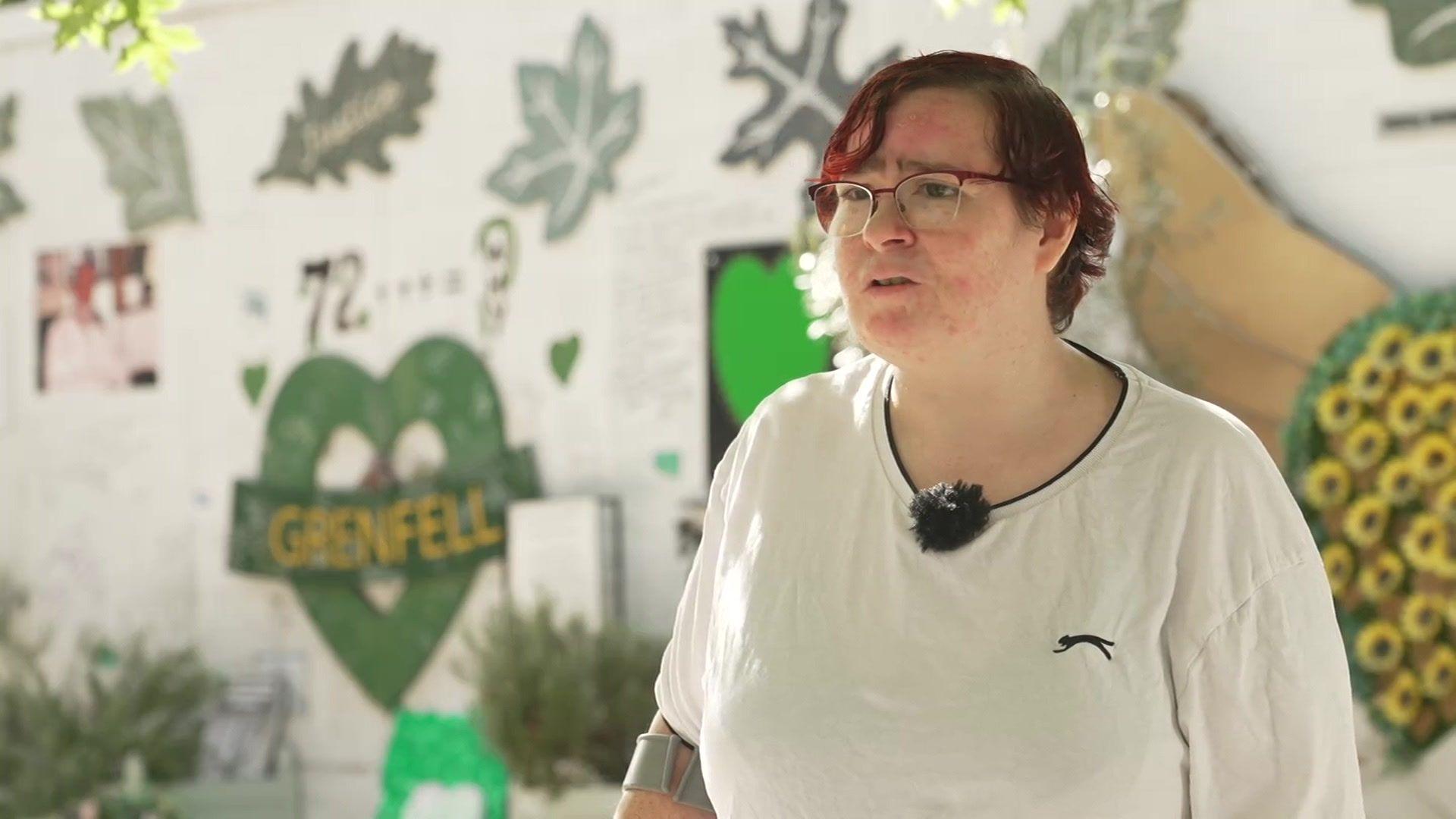 A person wearing glasses and a white T-shirt stands in front of a Grenfell memorial wall featuring green hearts, the number 72 and floral tributes.