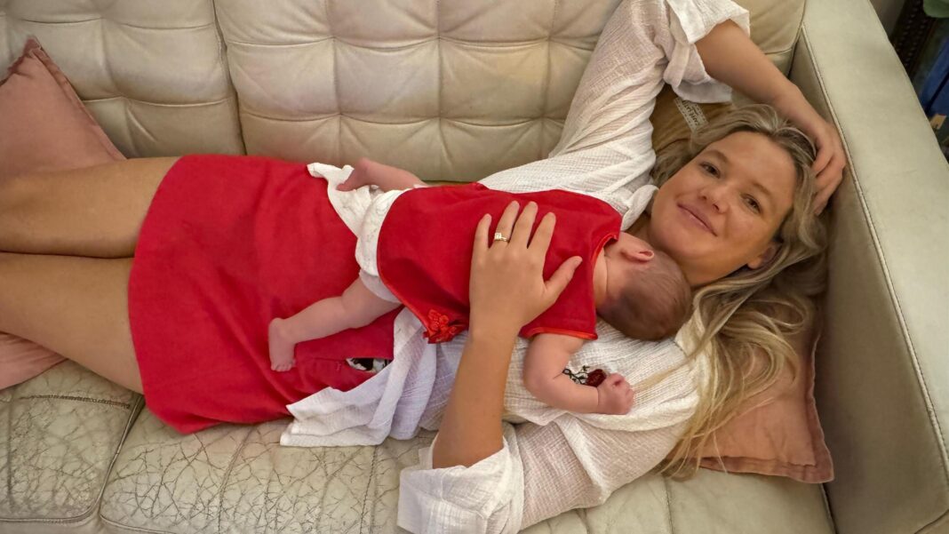 Deadly shark attack prompts rare kill order in Western Australia A woman with long blonde hair, wearing a white top and red skirt lying on a cream couch, with a baby sleeping on her chest.