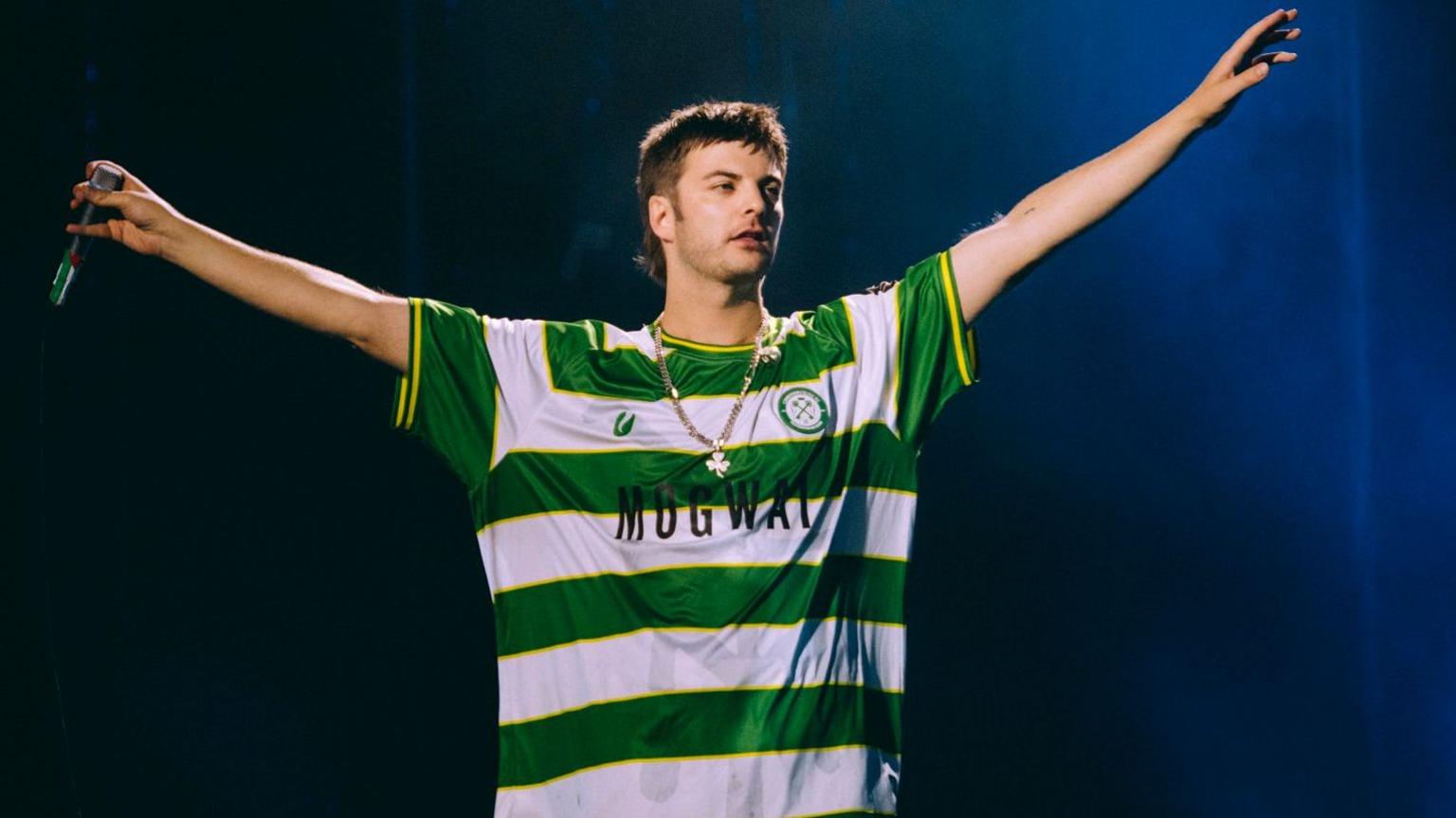 Grian Chatten performing at Reading Festival wearing a Cwmbran Celtic FC away shirt. He is looking away from the camera, with a microphone in one hand and the other raised in the air. The football shirt is green and white.