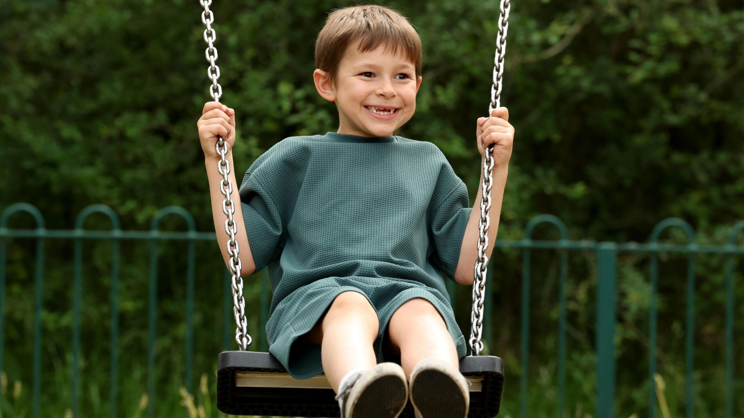 Elliot, a seven-year-old boy pictured on a swing outside. He has short brown hair and is wearing a green top and shorts. He is looking to the side and smiling. He has gaps in his teeth.