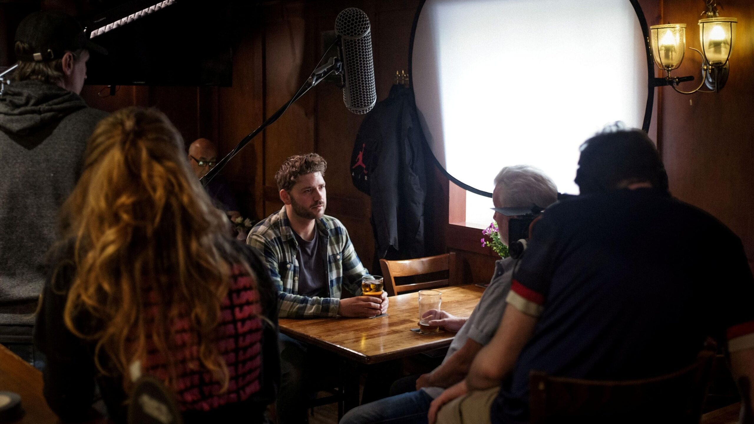 A behind-the-scenes view of a film or video production inside a wood-panelled pub, as  a man sits at a table holding a drink while crew members and equipment surround the scene.