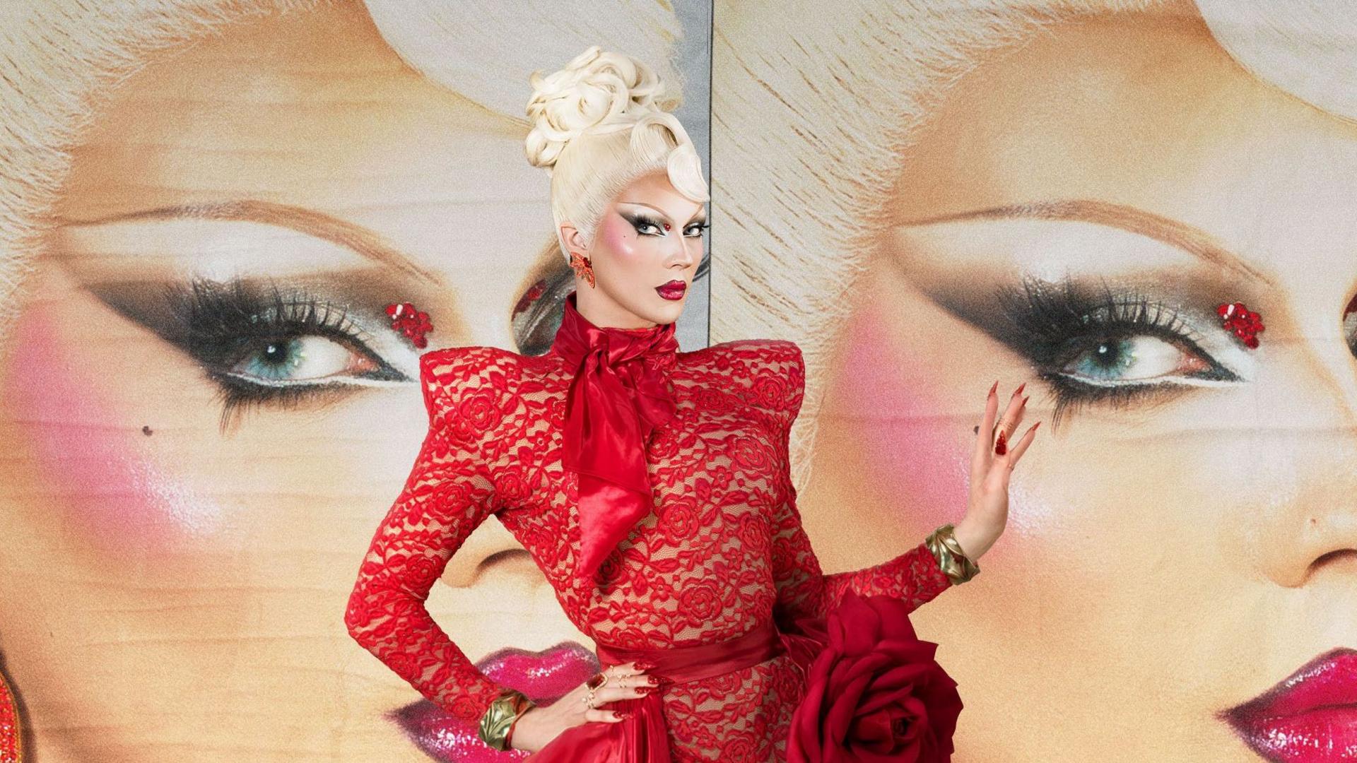 Naya Thorn wears a red lace dress and has a blonde wig on done in an up-do. She has dramatic dark eye makeup. She is standing in front of a poster board which has her face on it.