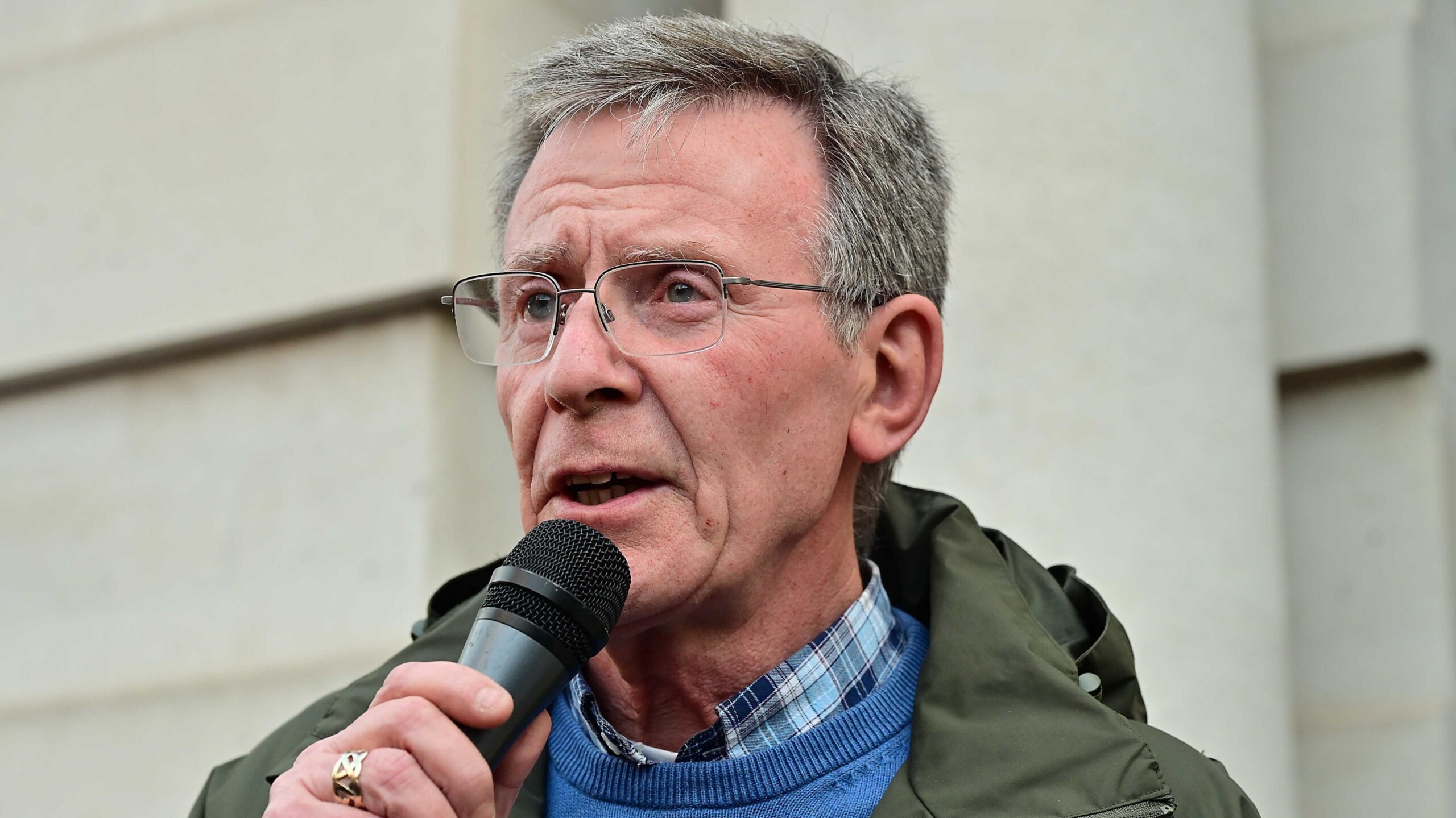A man is speaking. He's holding a mic. He has glasses on and is wearing a ring and a green coat with a blue jumper and a checked shirt.