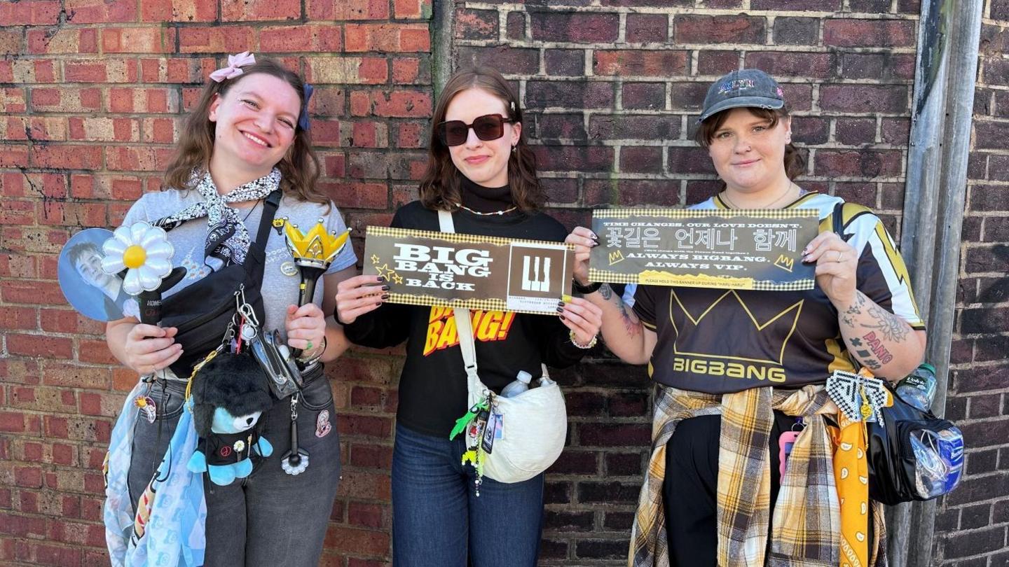 Three women standing in front of a brick wall display concert merchandise and banners. One holds a flower-shaped light stick and a crown-shaped light stick, with a cross-body bag, keyrings and a blue shopping bag. The centre woman holds a banner reading “BIG BANG IS BACK” and carries a white shoulder bag. The third holds a banner with Korean and English text and wears a clear waist bag. Various accessories, badges and merchandise are attached to bags and clothing.
