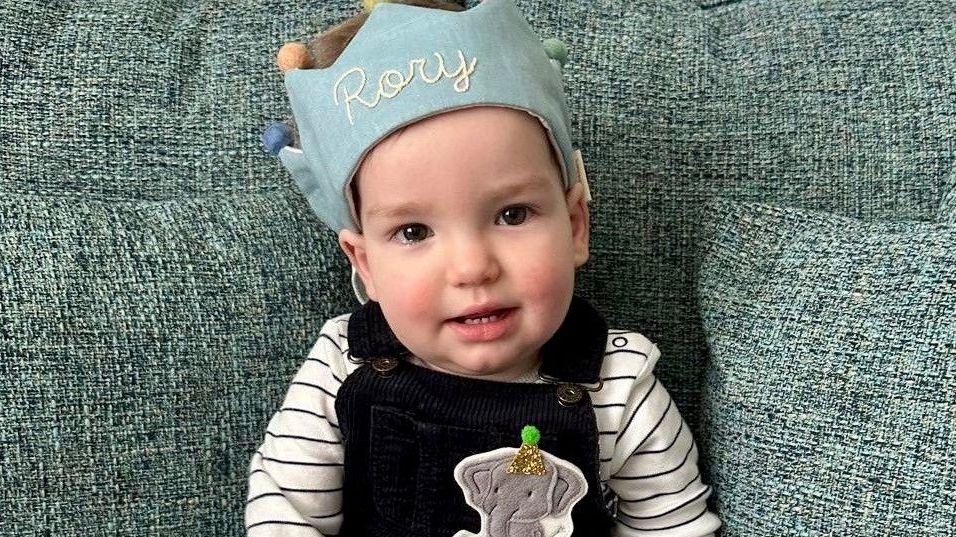 A one-year-old boy sat on a sofa with a crown saying "Rory" and he is wearing black dungarees with an elephant on.