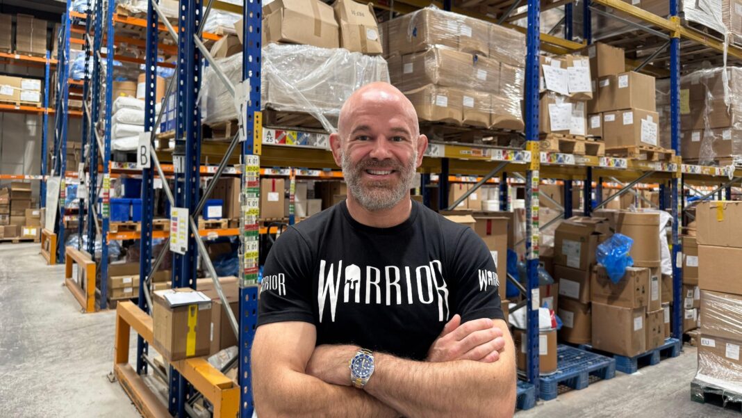 ‘Proteinflation’: Meet the people paying up to £100 for a bag of protein Kieran Fisher, a bald man with a short grey beard wearing a black T-shirt with the word WARRIOR on the front, standing smiling in a warehouse. Behind him are cardboard boxes stacked on blue metal shelves.