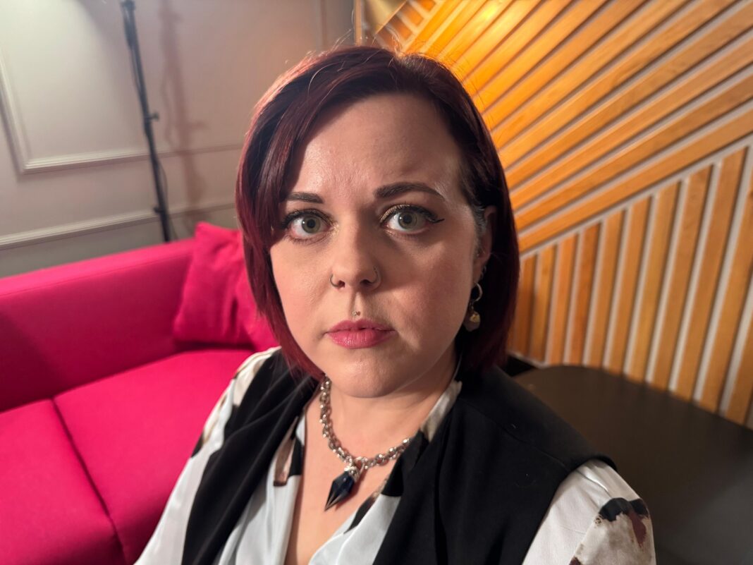 Headshot of Claire Milne, who has short dark hair, nose piercings and a piercing just above upper lip. She looks straight at the camera with a pensive expression on her face.