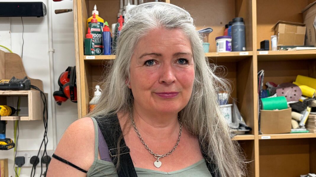 An older woman with long silver hair. She is standing in a workshop.