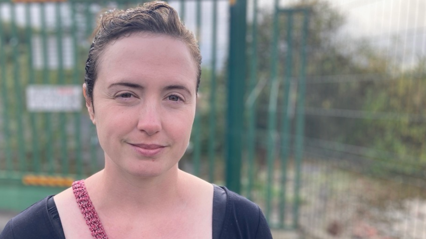 Natalie Dugdale is a white lady with brown hair wearing a blue t-shirt and a red handbag in front of a wooded area