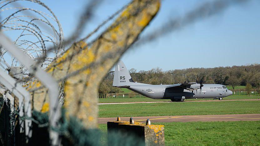 A large grey bomber parked on the runway at RAF Fairford on a sunny day, visible through the barbed wire of a metal perimeter fence. The grass is bright green and there are lots of trees in the distance.