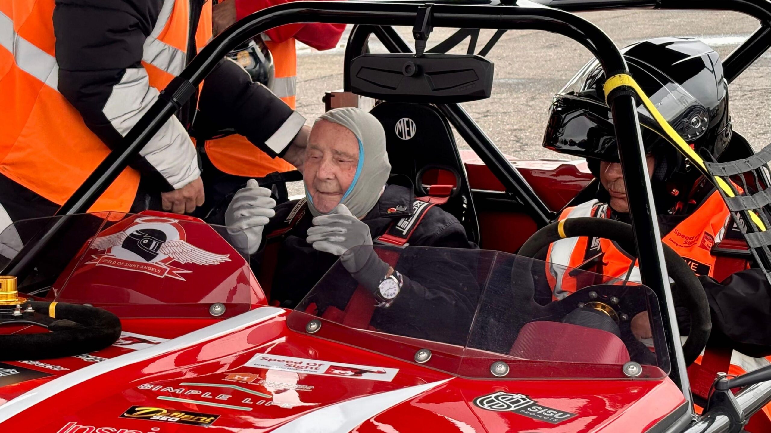 An elderly man wearing a balaclava is sitting in a red sports car.  He is smiling. Next to him is a driver wearing a helmet and hi vis vest.