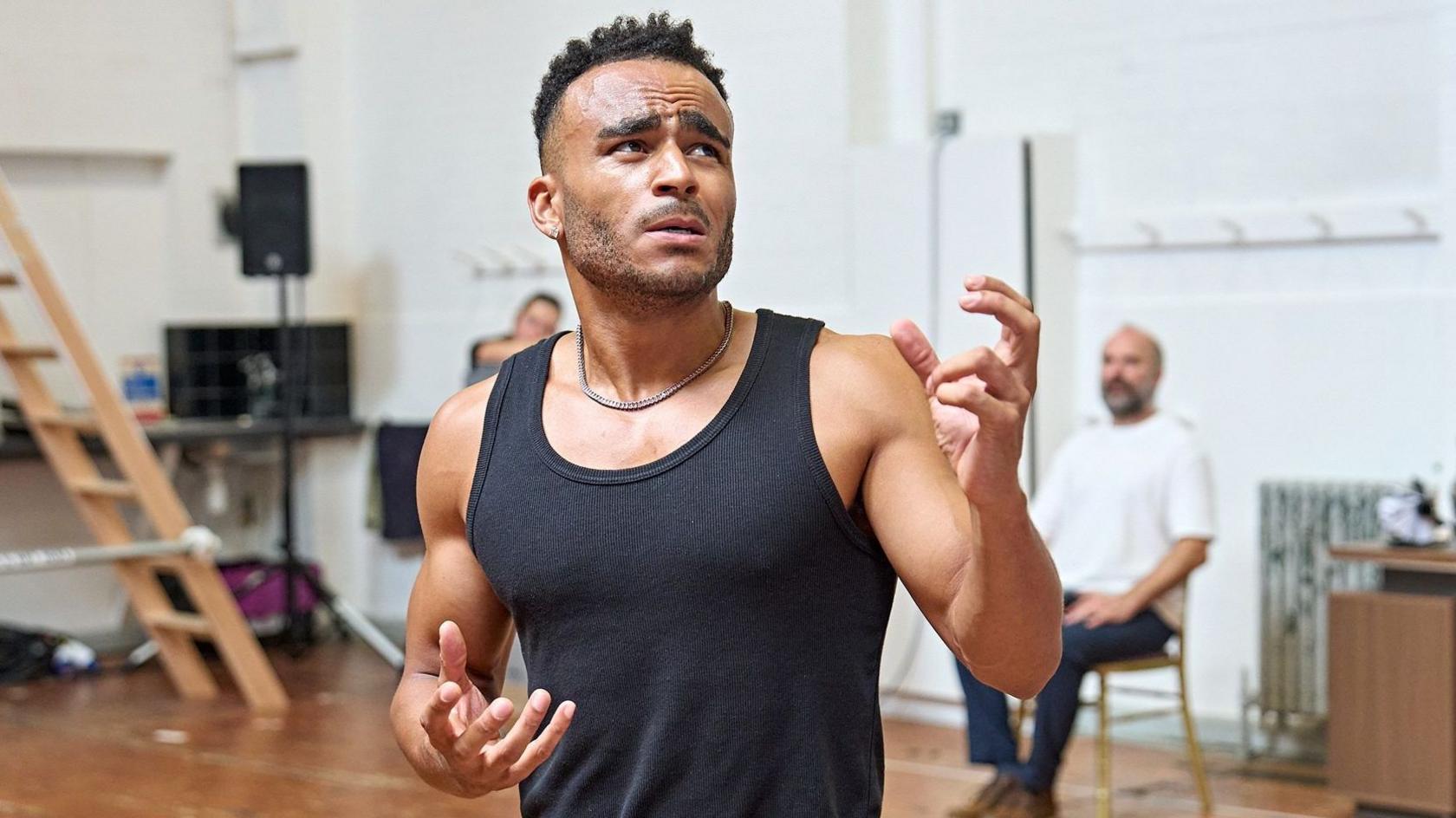 Munya Chawawa in a black vest with both arms gesturing and his face with a pained expression, in a rehearsal room