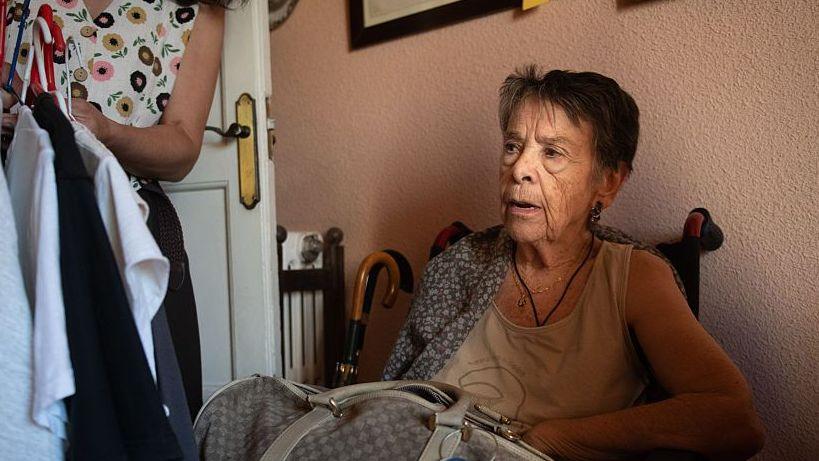 Maricarmen, a frail elderly woman, packs a bag with her belongings as she sits in a chair by the door of her flat