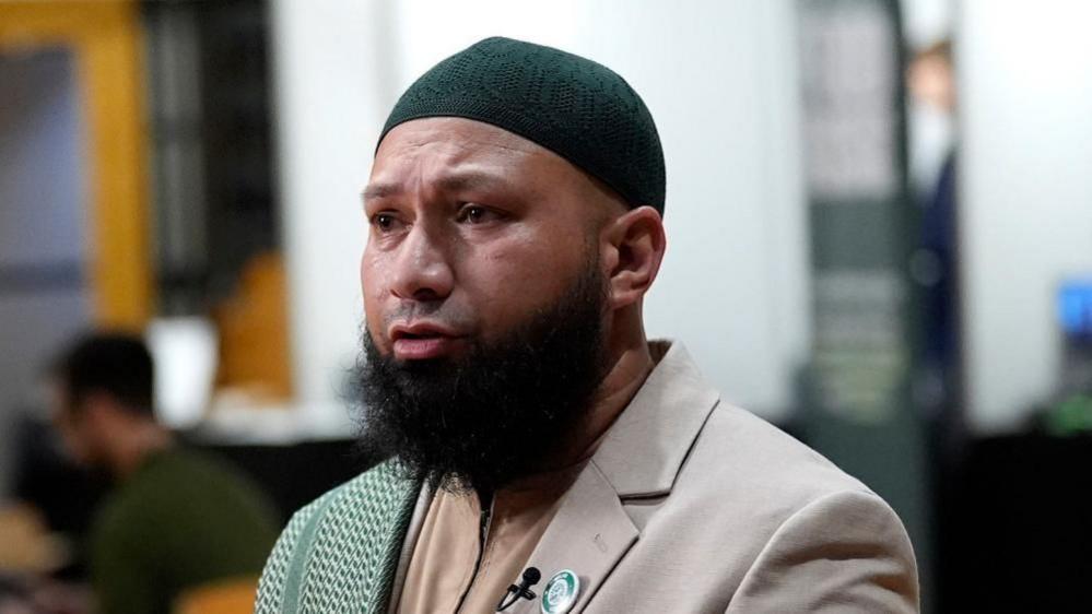 Mothin Ali looking to his right, wearing a beige suit jacket with a green and white scarf draped over his right shoulder
