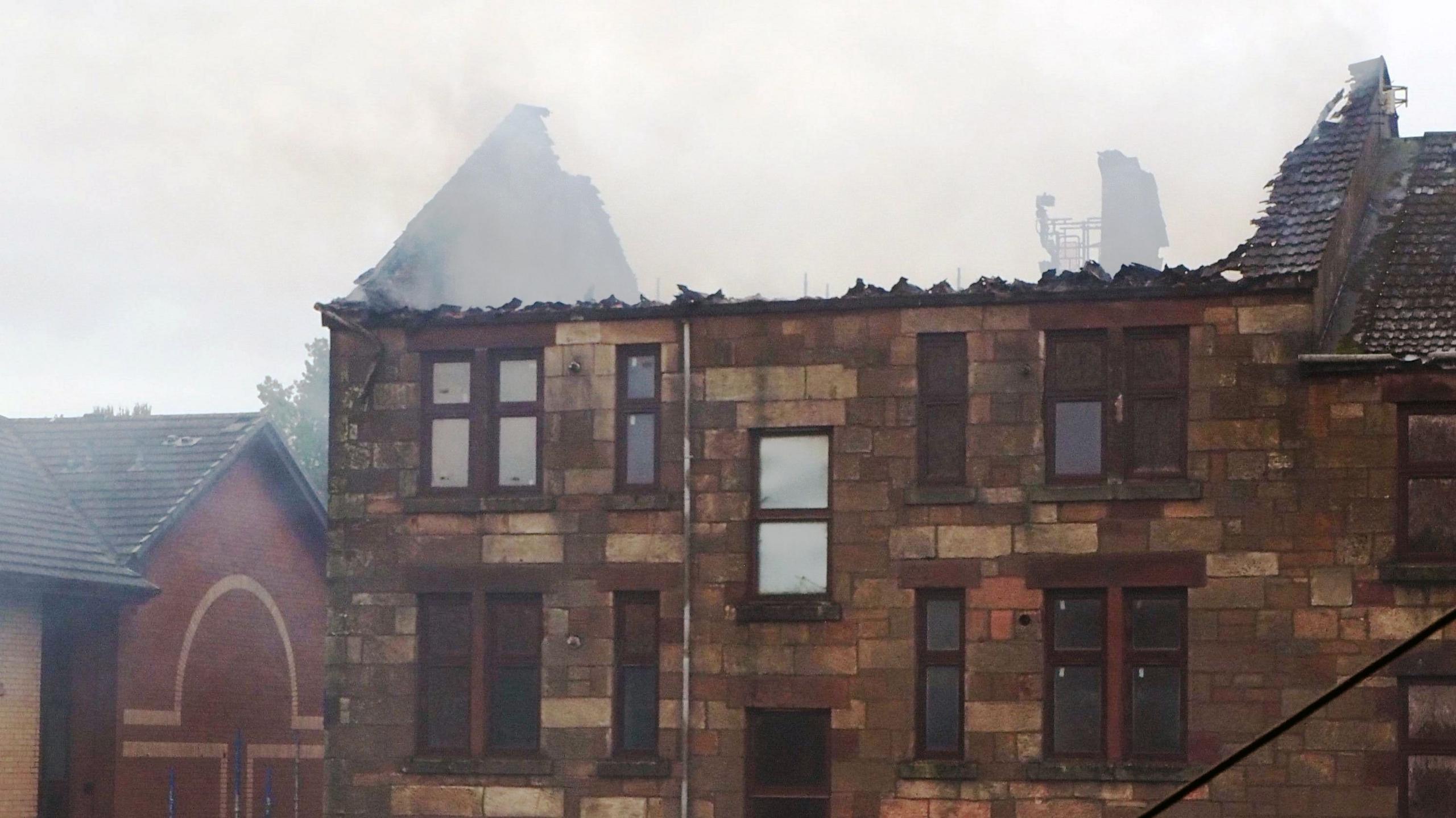 Burned-out tenement building which no longer has a roof. Three firefighters can be seen beside the building on the steps of a nearby flat
