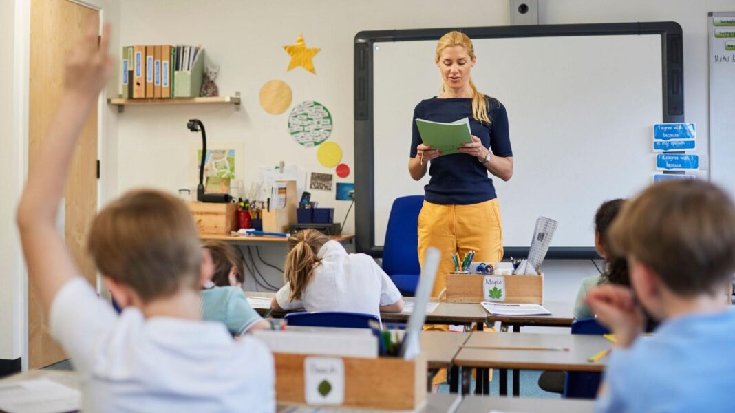Stock image of a teacher teaching her class. She is wearing yellow trousers and a navy blue long-sleeved tshirt. Behind her is a Smart board and classroom items. In front of her are children learning at their desks - one boy has his hand up.
