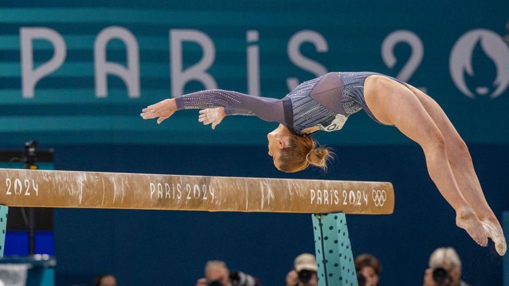 Alice Kinsella leaps backwards with her arms outstretched as she dismounts the balance beam at the Olympic Games in Paris. The beam has the word Paris on in white letters with the date of 2024 and the five Olympic rings logo.