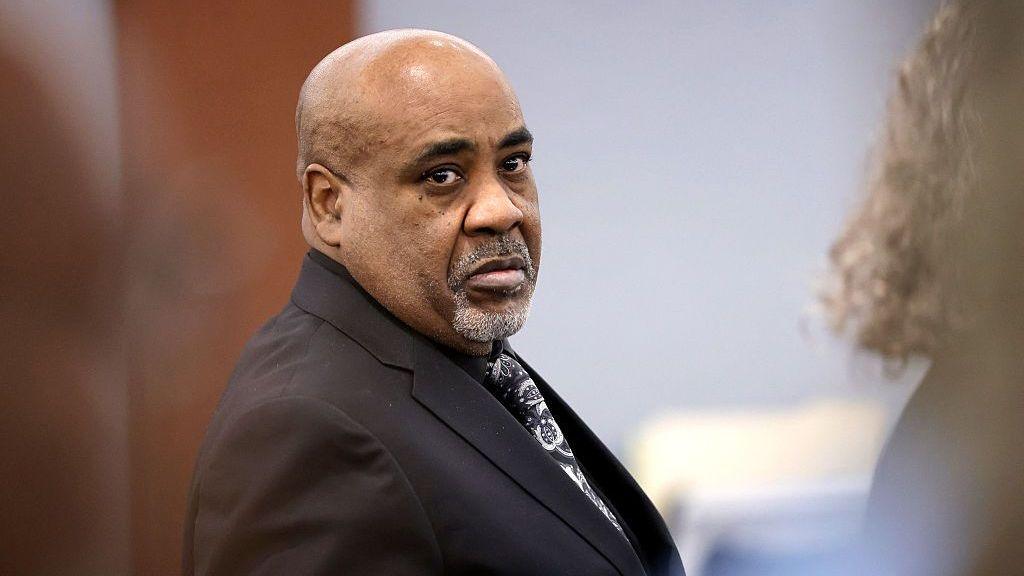 A man wears a black suit and patterned tie. He has a white goatee and serious expression.