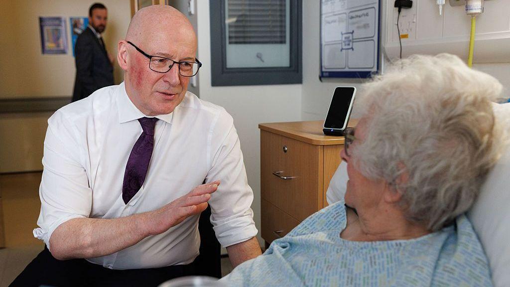 Swinney suggests Scots will not travel further for health care under reforms John Swinney talking to a female hospital patient who is in bed.
