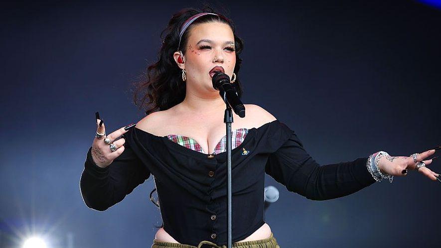 Lola Young performing on stage in August. She wears and off the shoulder black corseted top with a red and white tartan bralet peeping over the top. She wears her long dark hair loose, pushed back with a headband. She wears her signature statement makeup, including a red lip, heavy blush and gems on her cheekbones.