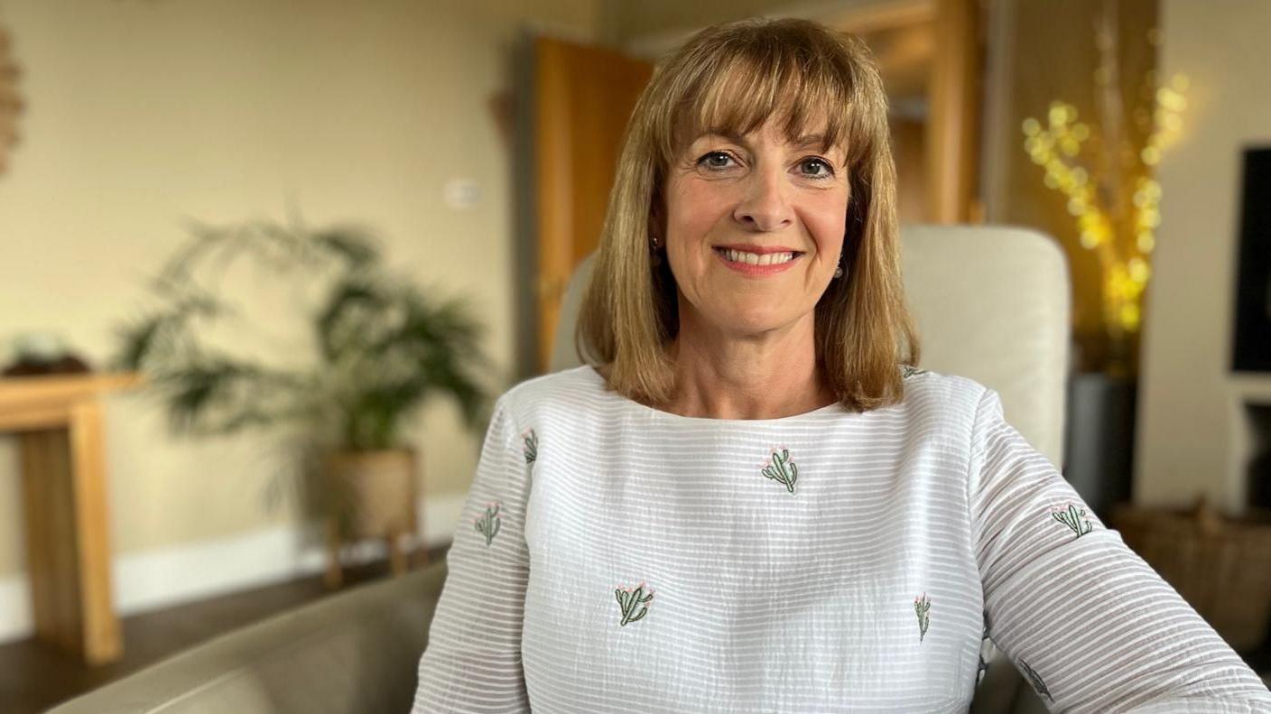 Emma is smiling at the camera, she is wearing a white blouse and is sat in a cream chair in a living room.