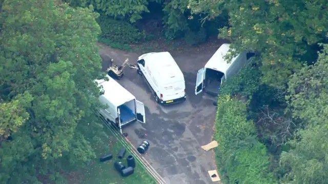 An aerial image showing three white vans with their rear doors open, parked on a fork road in a wooded area. A robot with a large mechanical arm is inspecting the front wheels of the middle van.