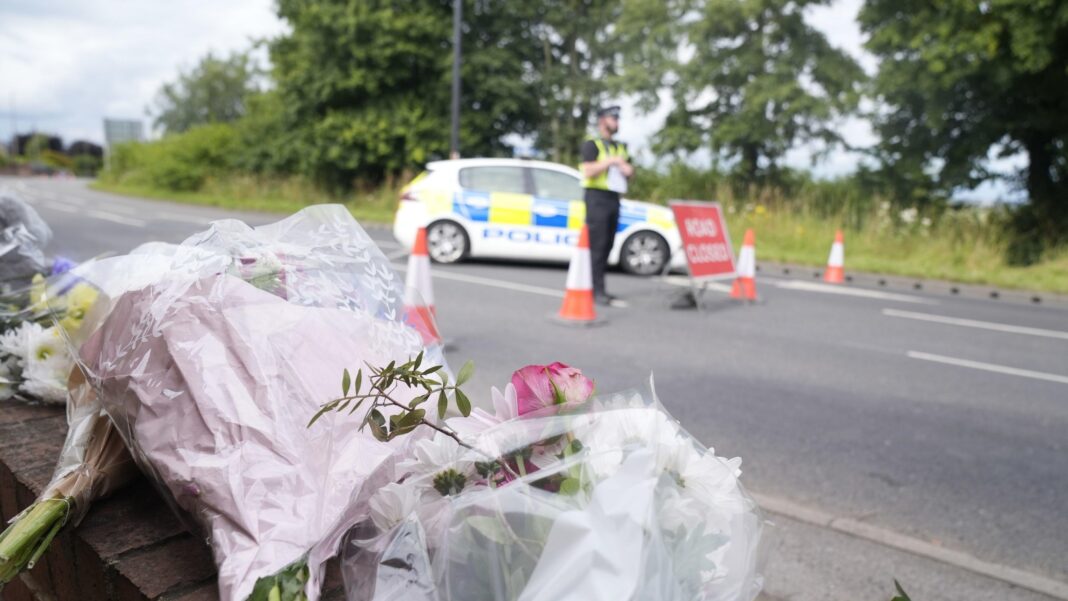 Several bouquets of flowers wrapped in plastic have been placed on a brick wall beside a road. In the background, a police car, traffic cones and a temporary sign reading “Road Closed” are positioned across the roadway, with an officer standing nearby. Trees and greenery line the road behind the police cordon under an overcast sky.