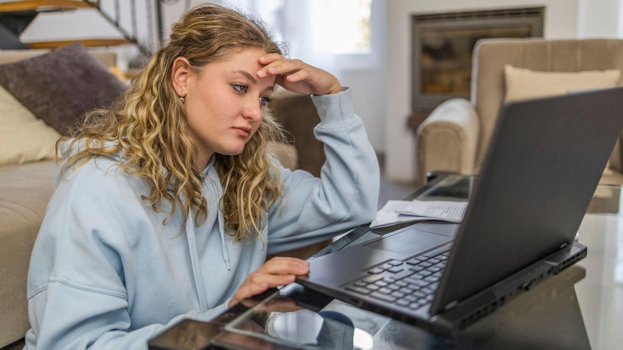 A teenaged girl who has long blonde curly hair and is wearing a light blue hoodie sits at a glass table, looking at an open laptop. One hand rests on the forehead while the other is near the keyboard. Papers lie beside the computer. She is in a living room with sofas, a staircase and a fireplace.