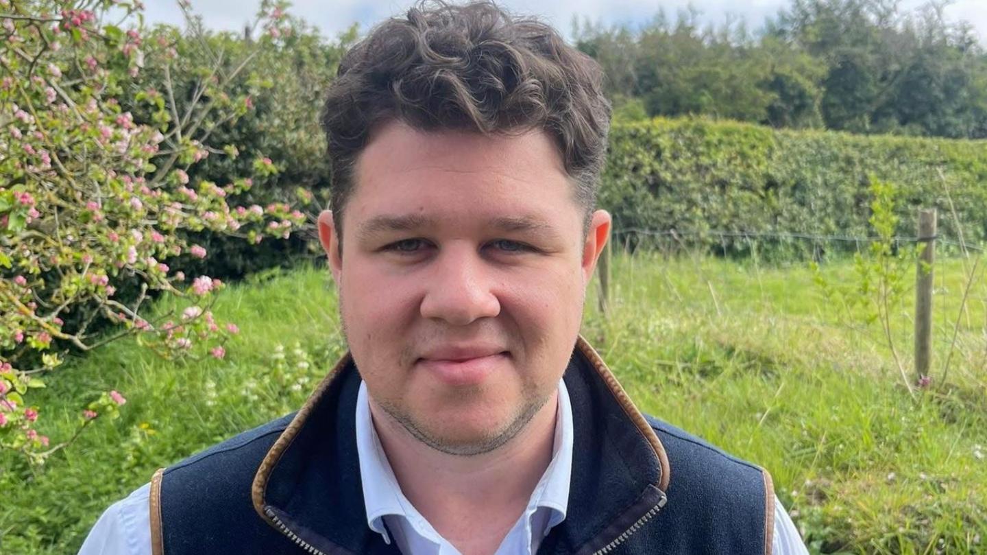 A man with dark hair smiles at the camera, wearing a dark blue gilet and a light blue shirt, in a field next to hedgerow