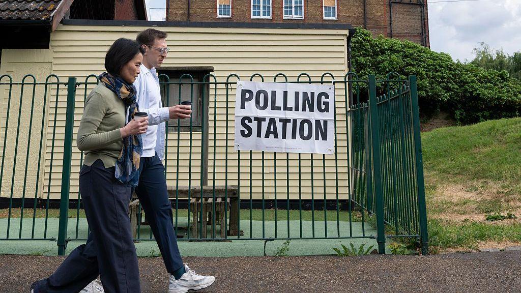 Two people walk past a polling station sign on a green fence.