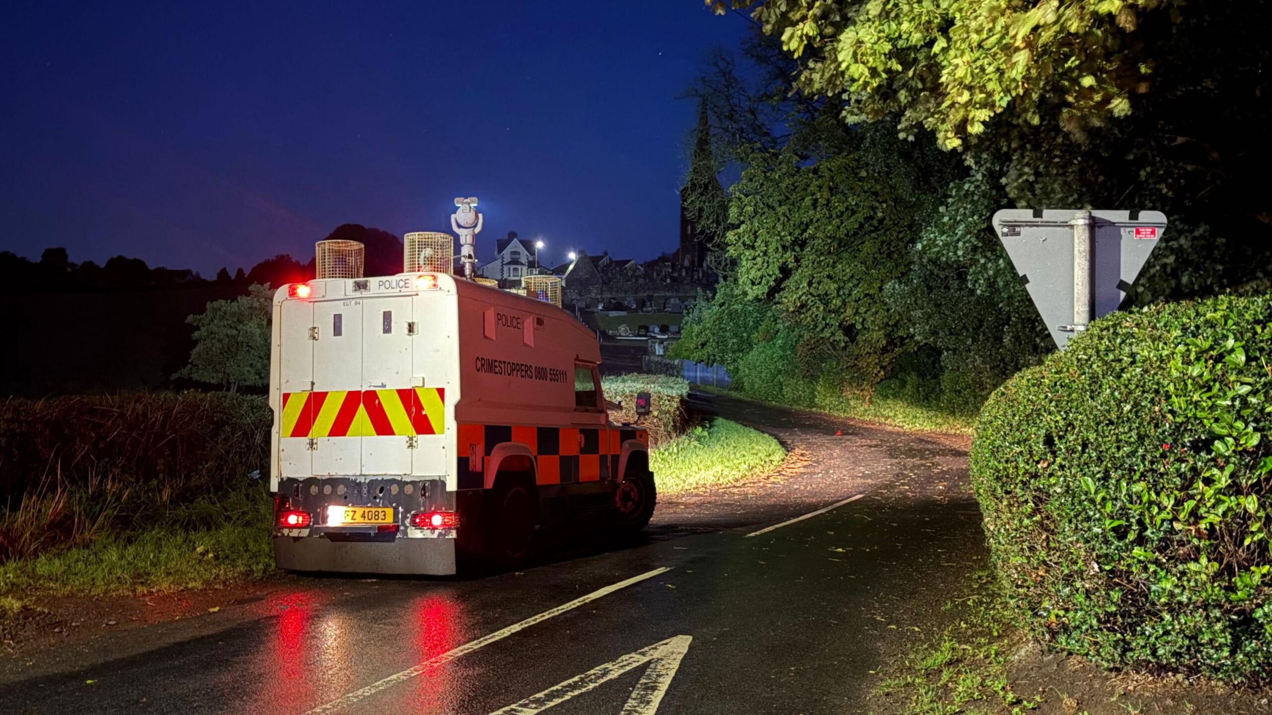 A police Land Rover sits on a rural road, it has yellow and red stripes and a blue an yellow checked pattern.