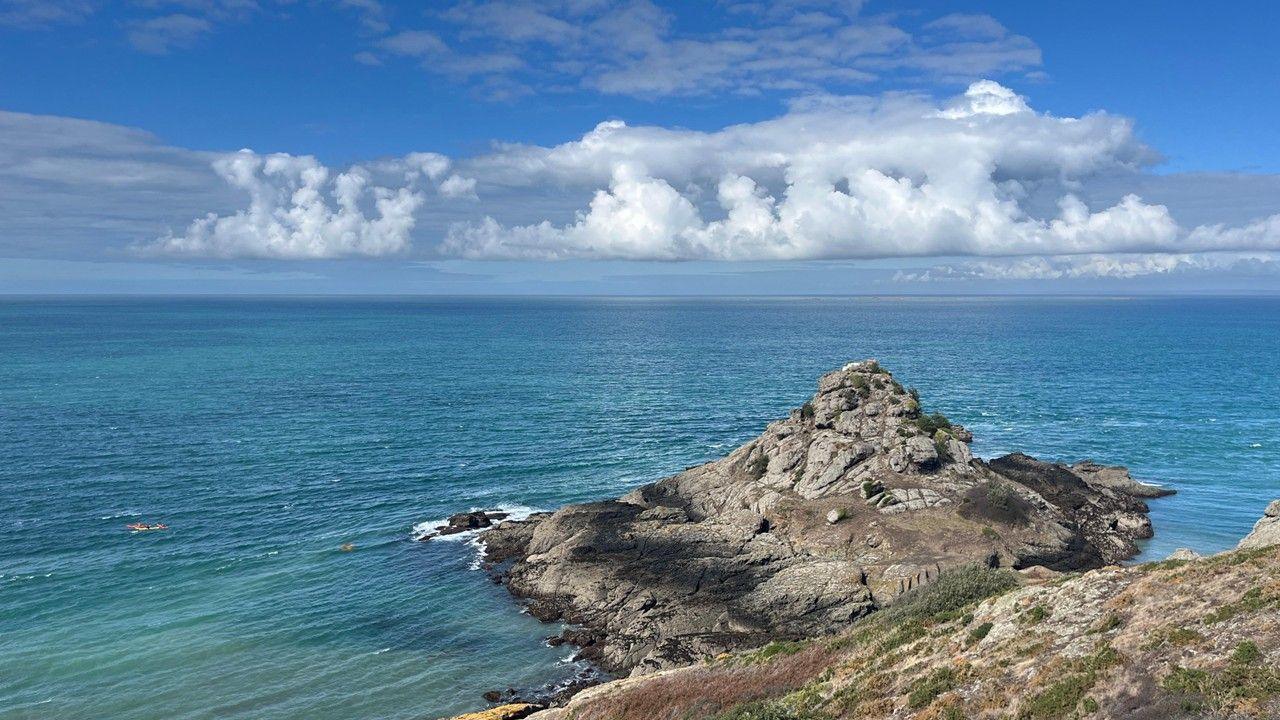 Blue sky with a long white cloud over blue green sea and rocks on the headland jutting out in the foreground.