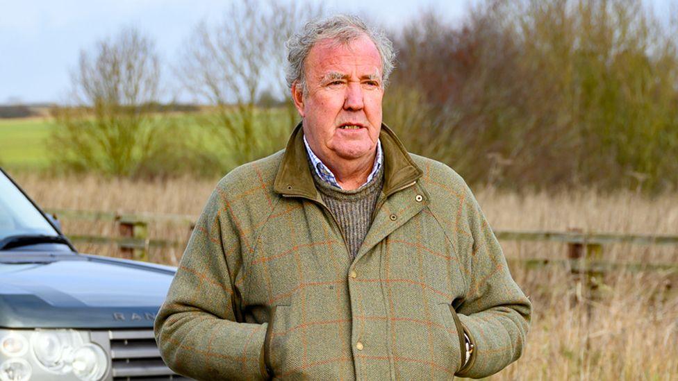 Jeremy Clarkson is pictured standing in front of a Range Rover car looking into the distance. He is wearing a green tweed coat and he has his hands in the pockets of the coat. There is a fence behind him and beyond that is a green field and some trees.