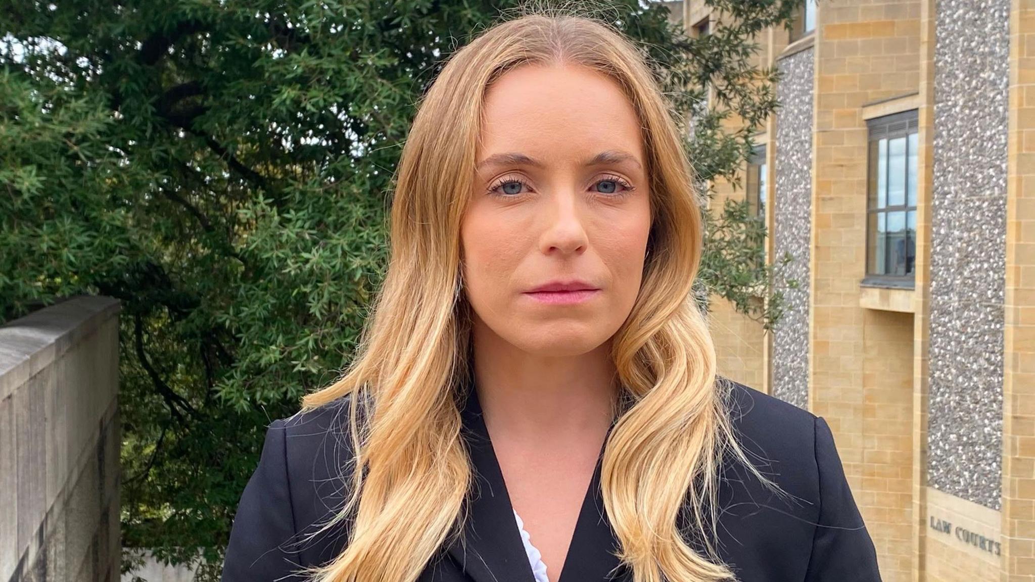 Adams looks at the camera with a serious expression. She has long blonde hair and is standing outside the court, with a large tree behind her.