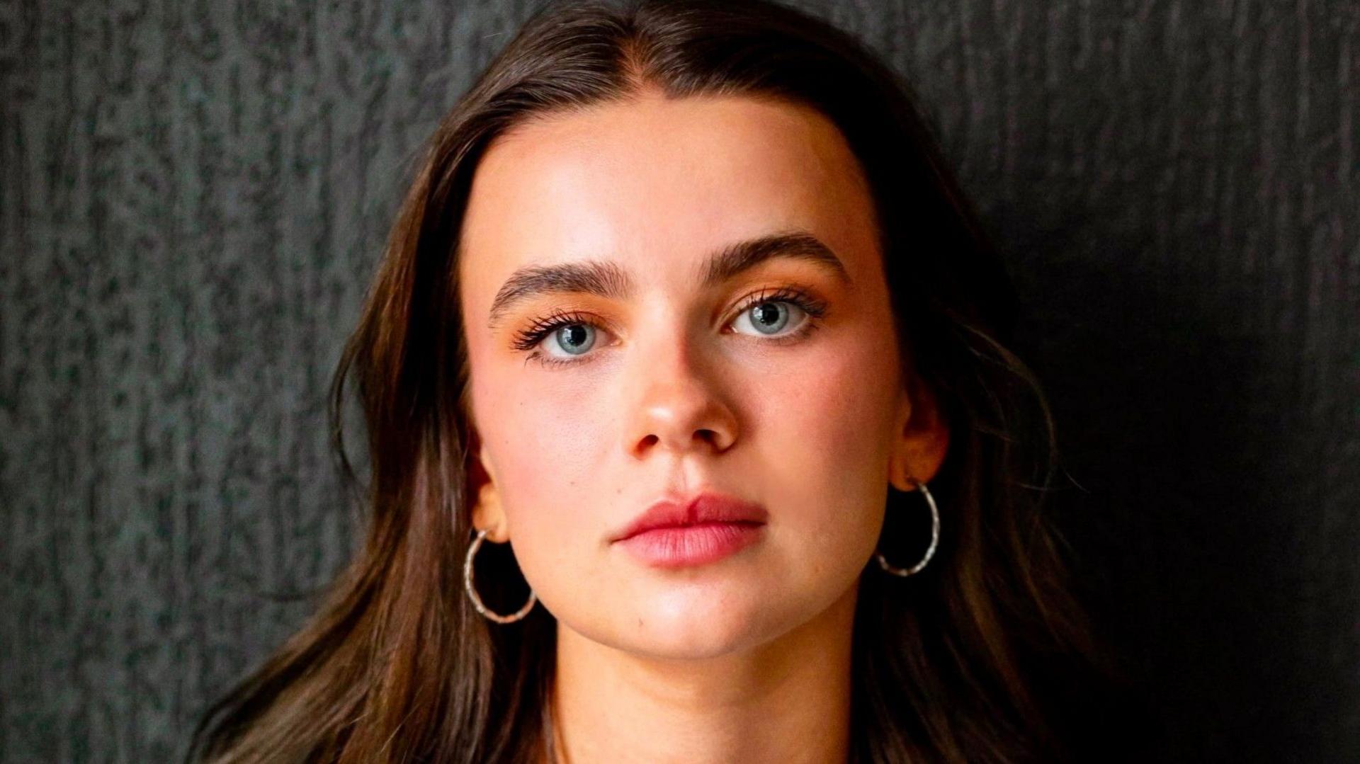 Tabitha Smyth has long brown hair looks to the camera as she wears hooped gold earrings. She has green eyes and stands in front of a black-grey wall.