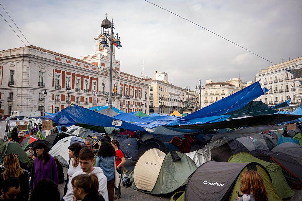 Blue tarpaulin hangs above dozens of tents in Madrid's Puerta del Sol. Protesters stand around them. A white and red brick building with a clock tower is in the background.