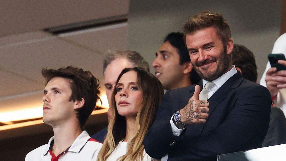 David Beckham alongside his wife Victoria and his son Cruz at a World Cup game, Beckham is wearing a suit and expressing a thumbs up.