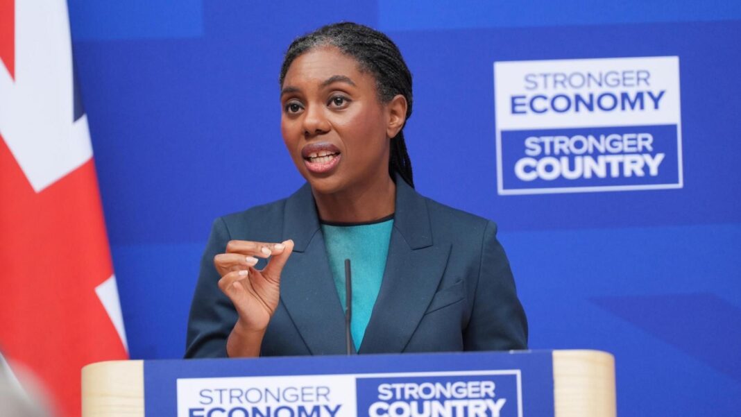 Badenoch accuses Burnham of kicking defence spending into the long grass Badenoch is pictured from below the podium where she is giving a speech, at London Scottish House in Westminster, with a royal blue background and a Union flag to her left. She wears her braided hair tied back in a low ponytail, with a navy jacket and a turquoise top.