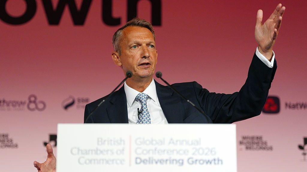Andy Haldane gestures animatically as he speaks from a white podium in front of a red background at a British Chambers of Commerce event.