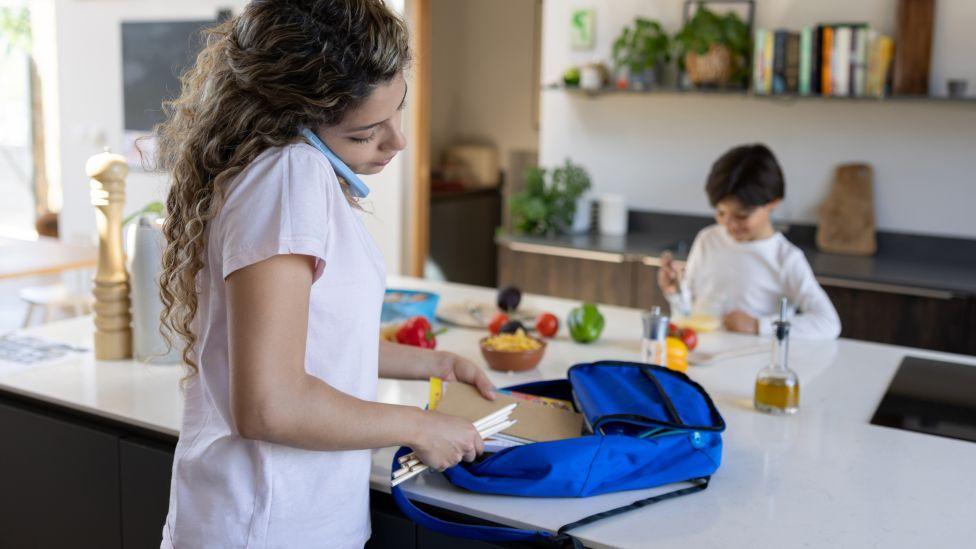 A woman with dark wavy hair packs pencils and notebooks into a blue rucksack in a kitchen, while a small children sat across from her eats breakfast