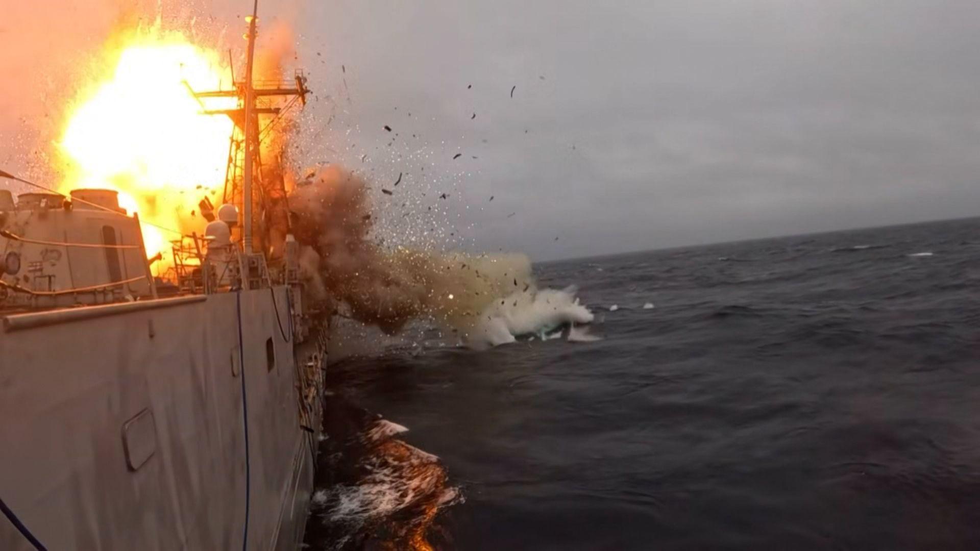 Orange flames and smokes erupts from an explosion on USS Klakring. Part of the grey warship is visible on the surface of a dark blue sea.