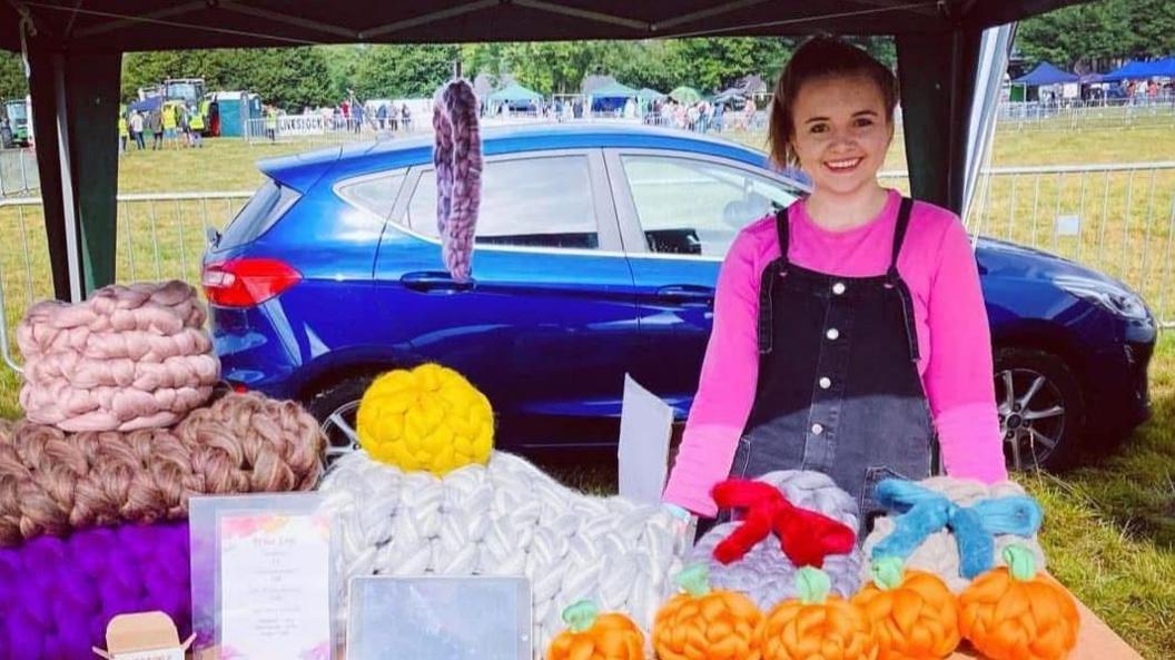 Harriet is stood in a field behind a stall with an open gazebo above her head. Several handmade wool products of various colours are on the table including five small knitted pumpkins and some large throws. In the distance are other trade stands, barriers, and people in hi-viz jackets. She is smiling and wearing a pink long-sleeved top under a blue denim dungaree dress, with her hair teid back in a ponytail.