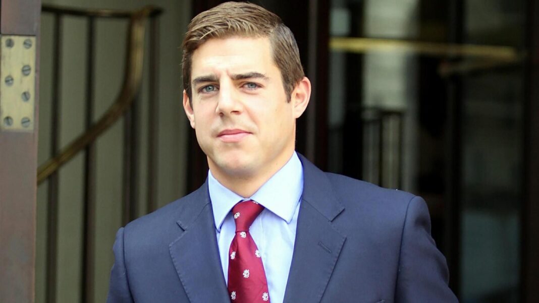 A head-and-shoulders image of Nicholas Brandram, seen with light brown hair and blue eyes, wearing a light blue collared shirt, a dark blue suit jacket, and a burgundy tie with a repeated white pattern. He is facing forward with a neutral expression.