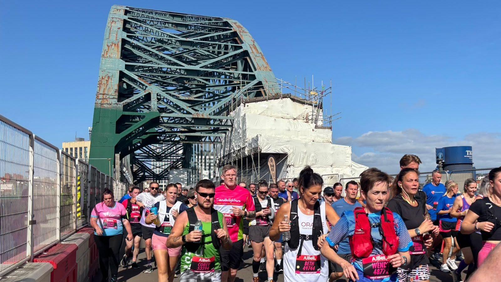 Runners in bright running gear stream across the green metal Tyne Bridge wich is obscured by scaffolding and bordered by metal fencing.