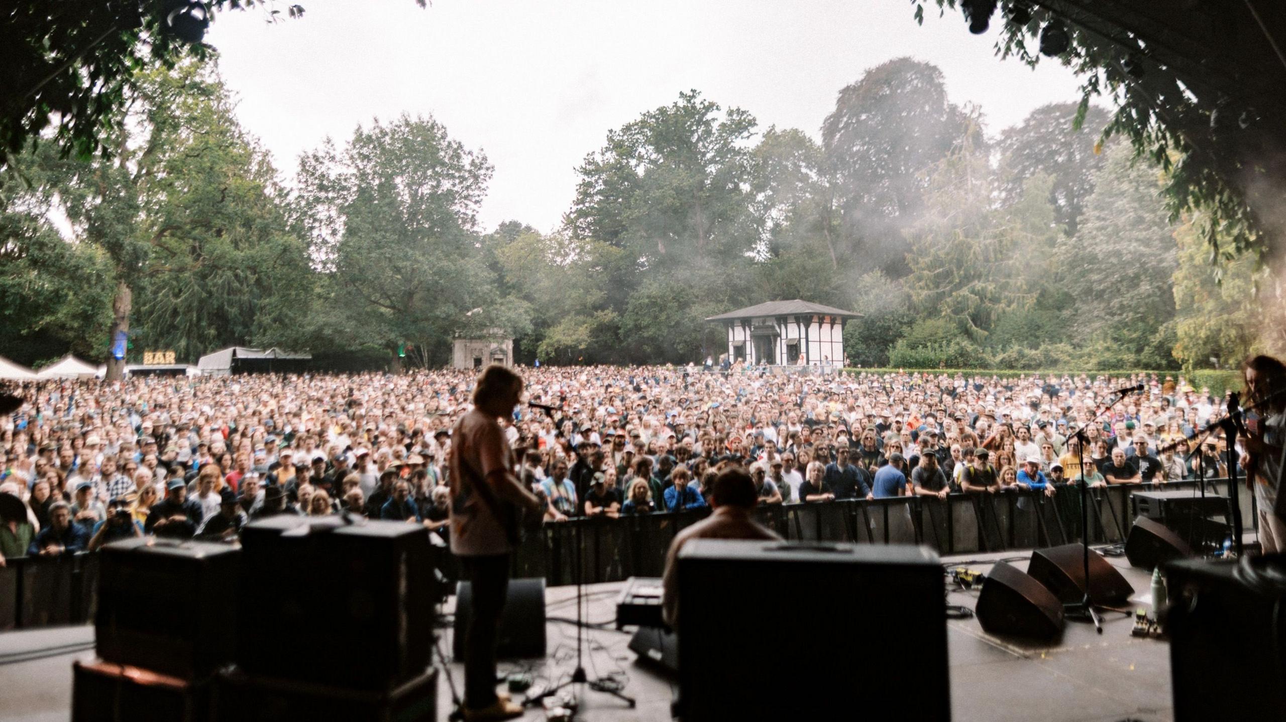 Crowds of people looking towards a stage with a musician stood on it and smoke in the air.