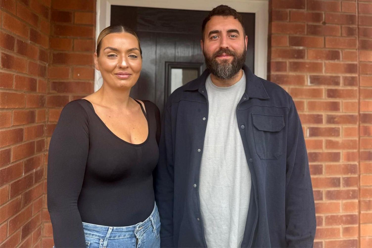 Jess and Darryl stand side by side at the entrance of a brick building, Jess in a black long‑sleeved top and jeans, Darryl in a light grey shirt and dark jacket, positioned in front of a dark door with a small window.