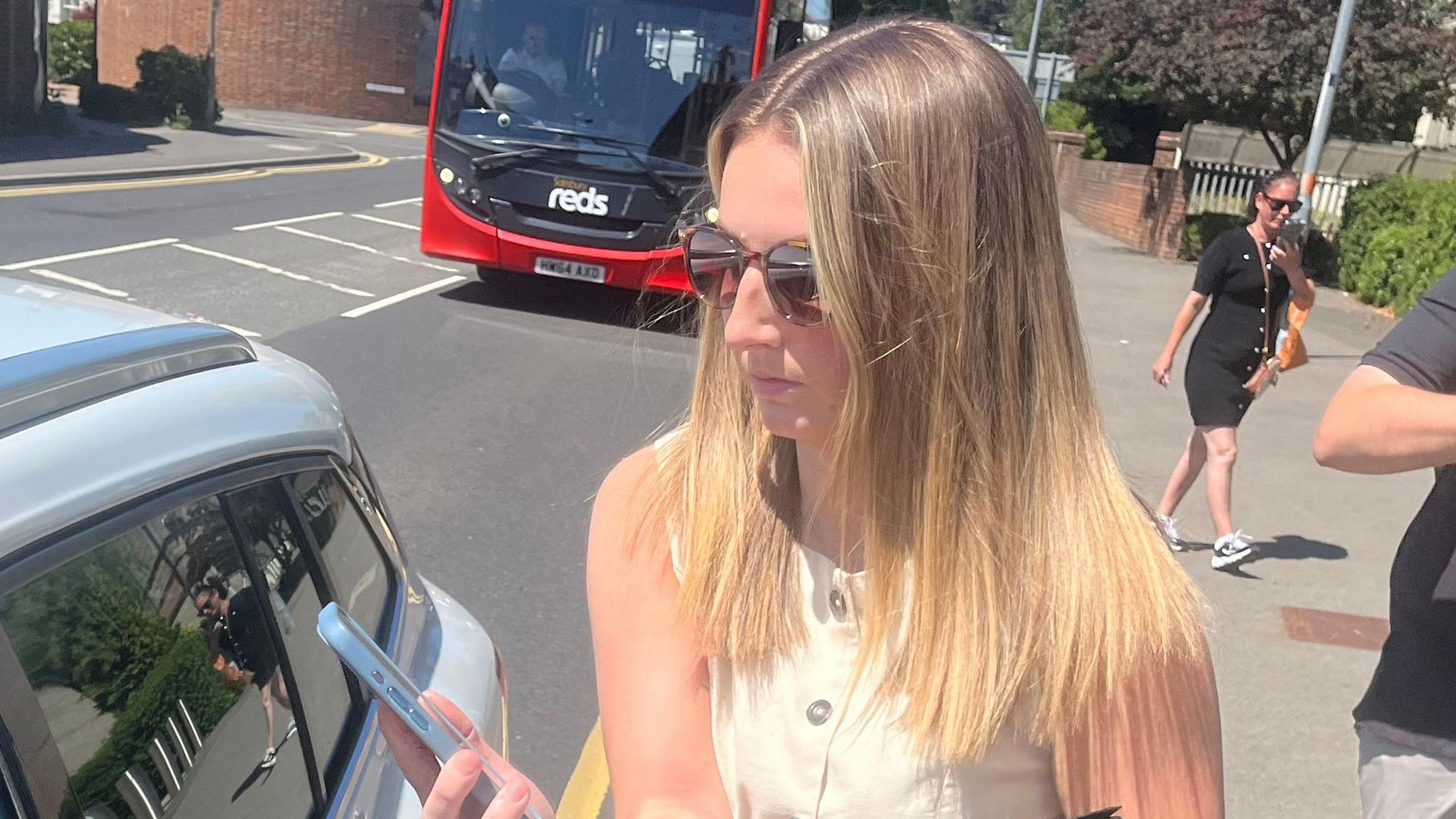 A woman with long blonde, straight hair is standing by a car looking at a blue mobile phone. She is wearing brown sunglasses and a beige waistcoat. There are people walking behind her and a red bus in the background.