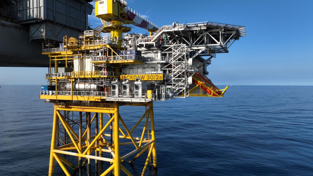 A mostly yellow-painted gas platform and allied grey rig stand in calm blue seas with a clear blue sky behind them. It is sunny.