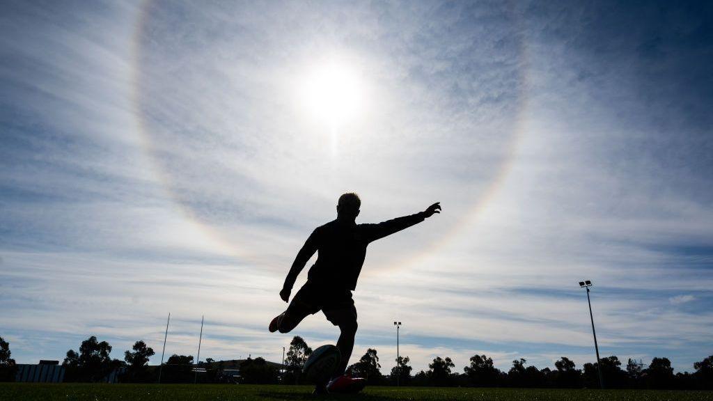 A silhouetted rugby player strikes the ball off a tee during a training session, with the sun glaring down from behind him