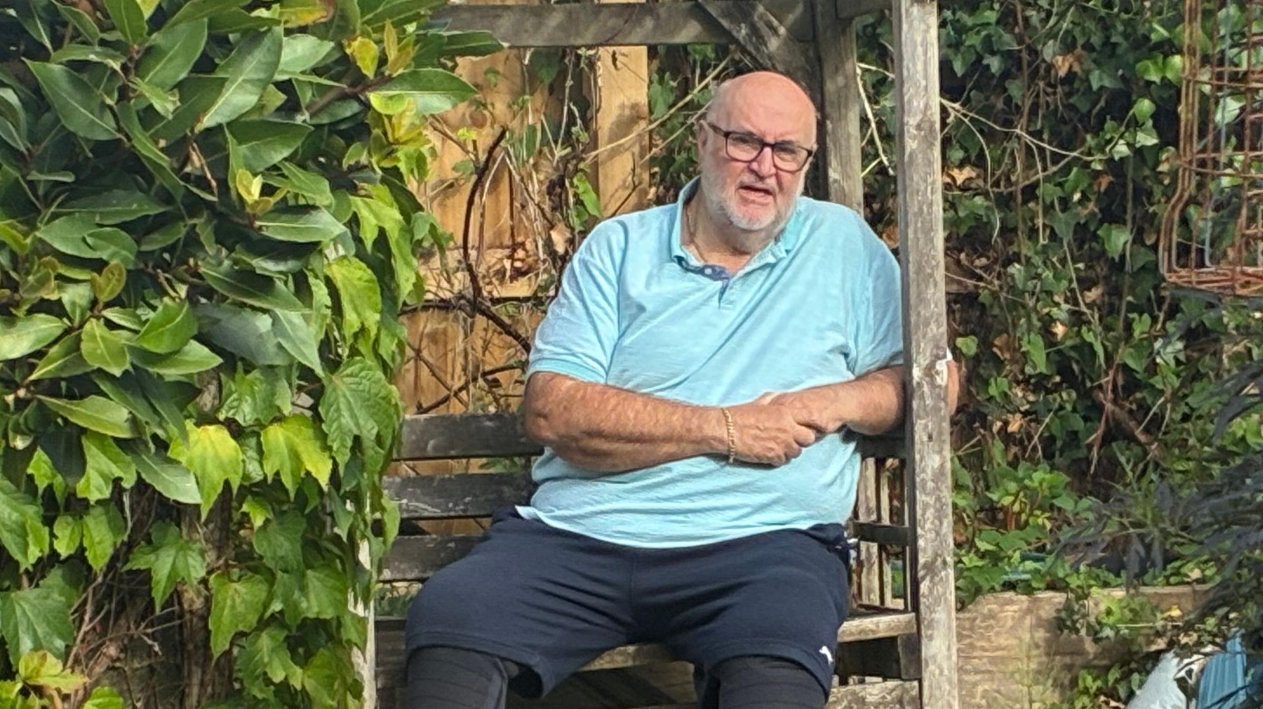 Phil Bond, 68 sat on a bench in his garden. He's wearing a light blue polo top and navy shorts, and slippers. He's bald with glasses. He's sat surrounded by greenery.